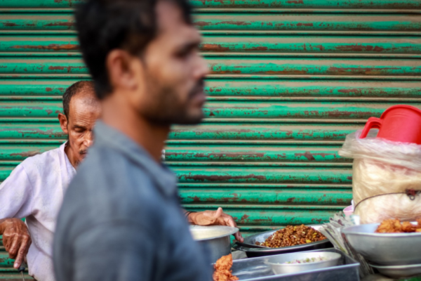 A street food vendor preparing food at a roadside stall, with another man passing in the foreground and a green metal shutter in the background.