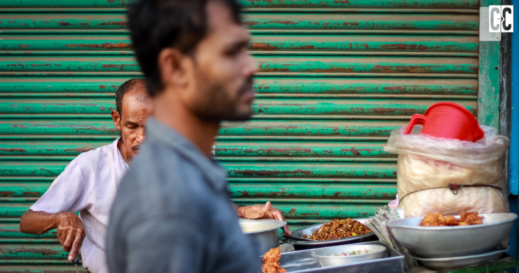 A street food vendor preparing food at a roadside stall, with another man passing in the foreground and a green metal shutter in the background.