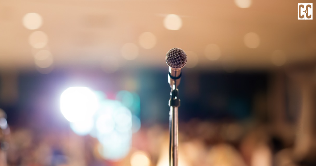 “A microphone on a stand against a softly blurred, warmly lit event venue.